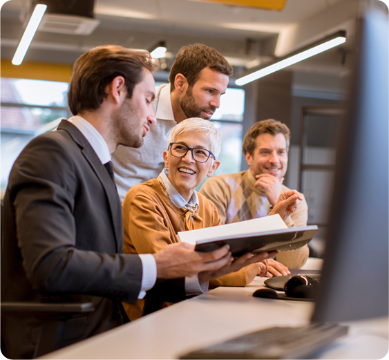 A woman in a yellow shirt speaks with a professionally dressed man holding a book, with two other men in the background, in an office environment.